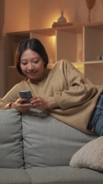 Dikey video. Çevrimiçi boş zaman. Mobil sohbet. Sosyal medya. Rahat, gülümseyen bir kadın telefon mesajı okuyor rahat bir odada koltukta oturuyor..