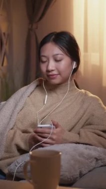 Dikey video. Müzik rahat. Huzurlu bir kadın. Ev tatili. Rahatlamış kadın dinlenme müzik listesi kulaklıkla evdeki koltukta telefonla dinleniyor..