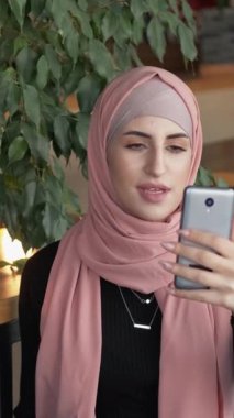 Dikey video. Çevrimiçi yayın. Video iletişimi. Tesettürlü neşeli kadın, takipçilerine el sallıyor. Sıcak kafede sosyal medya içerikli aboneleri selamlıyor..