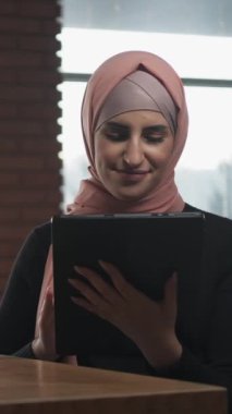 Dikey video. Online iş. Uzaktan kumanda. Keyifli kadın tesettürlü serbest yazar rahat kafede dijital tablet internette sohbet ediyor..