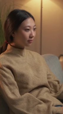 Dikey video. Mutlu insanlar. Ev tatili. Huzurlu bir şekilde gülümseyen bir kadın modern oturma odasında tek başına oturuyor..