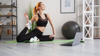 Germe egzersizi. Online oturum. Jimnastik dersi. Kas ağrısı giderici. Serbest dolaşım. Spor giyim bölümünde kadın fitness eğitmeni. Yoga minderinde bacağını çekiyor. İç dizüstü bilgisayarı da evde..