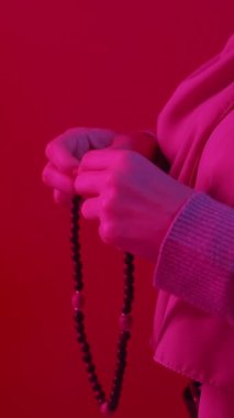 Dikey video. Tahta tespih. Ramazan namazı. Kırmızı fotokopi arkaplanda elinde tasbih boncuklarıyla dua eden tanınmayan bir kadın..