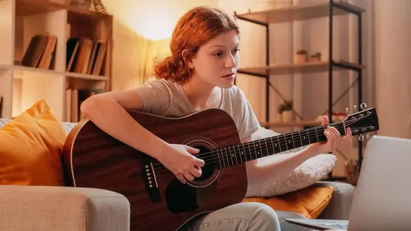 stock image Guitar learning. Internet music class. Inspired curious woman playing acoustic instrument watching video lesson on laptop at home.