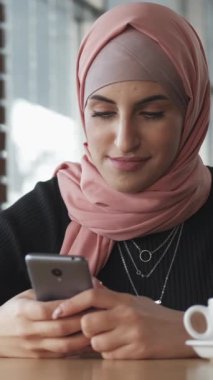 Dikey video. Cep telefonu kullanıcısı. Kahve keyfi. Kafe penceresindeki internet uygulamasında akıllı telefondan sohbet mesajı yazan tesettürlü bir kadın..