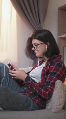 Dikey video. Ağ araştırması. Ev tatili. Kulaklıklı mutlu kadın akıllı telefondan arkadaşına mesaj atıyor. Oturma odasında dinleniyor..