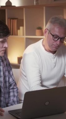 Dikey video. Ödevlerinizi birlikte yapın. Baba yardım et. Aile eğlencesi. Mutlu baba, laptopun başında neşeli oğluyla ders tartışıyor..