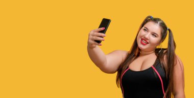 Fotoğraf çekiyorum. Sosyal medya. Video blogu. Aşırı kilolu komik bir kadın modeli selfie çekiyor. Sarı boş alan arka planında izole edilmiş telefon akışı kullanıyor..