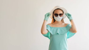 Kapalı tatil. Yolculuk hazır. Tıbbi maske takan kadın eldivenleri giyip hasır şapkaya dokunuyor ve boş beyaz arka planda izole bir şekilde duruyor..