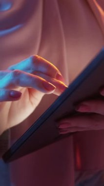 Dikey video. Sosyal ağ. İnternet araştırması. Dijital cihaz. Tanımlanamayan kadın elleri renkli neon ışığı altında bilgisayar tabletinde çevrimiçi web servisini tarıyor.