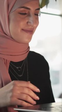 Dikey video. Laptop çağrısı. Çevrimiçi iletişim. Kafede oturmuş sohbet eden tesettürlü neşeli, arkadaş canlısı bir kadın..