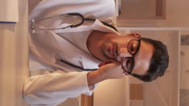 Dikey video. Yorgun doktor. Klinik aşırı çalışma. Hastane üniformalı, yorgun, profesyonel bir adam iş yerindeki çalışma masasında uyuyor..