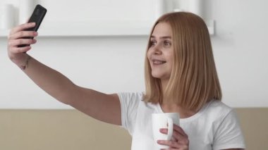 Medya çatışması. Çevrimiçi yaratıcı. Neşeli genç bayan blogcu telefonla konuşuyor video kaydediyor gülümsüyor parlak odada elinde kupayla gülüyor.