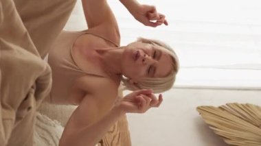 Dikey video. Sabah şarkısı. Hayatın tadını çıkarıyorum. Kaygısız, mutlu, iyi dinlenmiş, uyku tulumuyla dans eden, hafif yatak odasında, iç mekanda, yatakta oturan bir kadın..