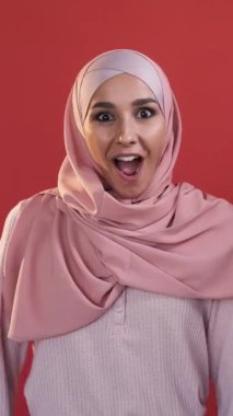 Dikey video. Şaşırmış yüz. Vay be sürpriz. Beklenmedik bir teklif. Heyecanlı, tatmin olmuş, ağzı açık, tesettürlü, kırmızı üzerine izole edilmiş bir kadın..