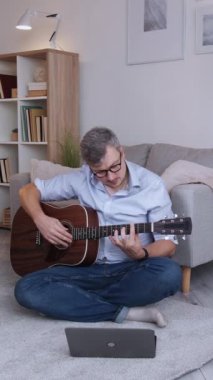 Dikey video. Evde eğitim. Müzisyen öğrenci. İnternetten ders. Akıllı konsantre adam, akustik gitar akorları üzerinde çalışıyor. Oturma odasının zemininde oturmuş, fotokopi çekiyor..