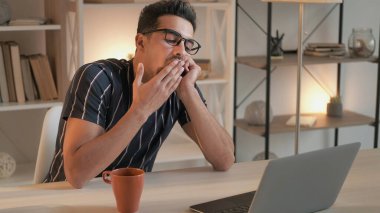 Uykulu öğrenci. Video dersi. Sıkıcı bir eğitim. İnternette dizüstü bilgisayarda ders veren yorgun genç bir adam. Masasında oturuyor. Oturma odasında iç mekânda esniyor..