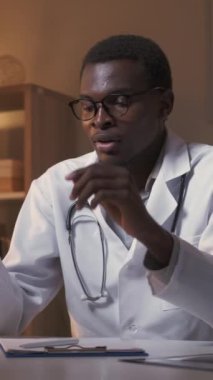 Dikey video. Yorgun doktor. Profesyonel yorgunluk. Ön cephede aşırı çalışma. Yorgun erkek doktor gözlüklerini çıkarıyor. Gözlerini ovuşturuyor, boynu esnetiyordu. Gece vardiyasında çalışıyordu..