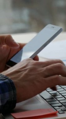Dikey video. İş teknolojisi. Dijital çalışma. Ofisteki adam cep telefonunda e-posta gönderiyor ve modern işyerindeki dizüstü bilgisayarında proje verileri yazıyor..