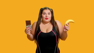 Seçenek yemek. Beslenme problemi. Kıvrımlı aşırı kilolu kadın, sarı arka planda izole edilmiş muz veya sağlıksız lezzetli süt çikolatası yediğinden şüpheleniyor..
