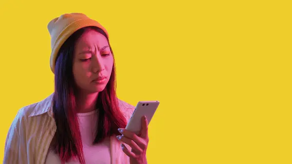 stock image Mobile fraud. Connection problem. Pink neon light confused frowning woman using phone isolated on yellow empty space background.