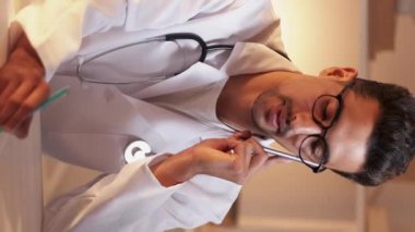 Dikey video. Gezici danışmanlık. Doktor telefon desteği. Tıp üniformalı zeki bir doktor, klinik masasında oturmuş, akıllı telefon ile tanı hakkında notlar yazıyor..