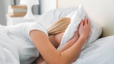 Uykusuzluk yorgunluğu. Sabah depresyonu. Tanımlanamayan, uykulu kız yastığa sarılıyor, migrenden muzdarip beyaz yatak odasında yüzünü saklıyor..