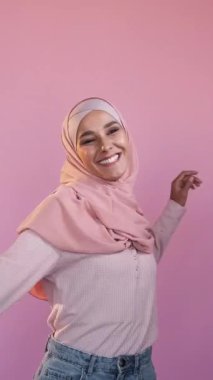 Dikey video. Mutlu danslar. Keyfin yerinde. Başarı kutlaması. Neşeli, iyimser, tesettürlü kadın pembe boş arkaplanda izole edilmiş eğleniyor..