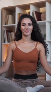 Dikey video. Evde yoga. Ev içi meditasyon. Sağlığın için. Rahatlamış, gülümseyen, nefes alıp veren, lotus bacaklı bir kadın oturma odasının içinde poz veriyor..