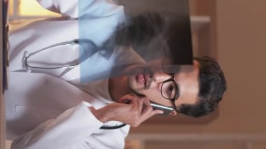 Dikey video. Telefon teşhisi. Gezici doktor konsültasyonu. Zatürree hastalığı. Akciğer röntgeni olan akciğer uzmanı akıllı telefonla konuşuyor. Kliniğin iç kısmında, çalışma masasında oturuyor..