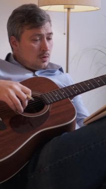 Dikey video. Beste yapıyorum. Müzisyen yaratıcılığı. Konsantre olmuş adam günlükte oturup akustik gitar çalıyor hafif ev iç mekanında yeni melodi akorları çalıyor..