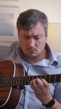 Dikey video. Melodi çalışması. Yaylı çalgılar. Yaratıcı hobi. Sıradan duygusal erkek sanatçı akustik gitar çalıyor evde keyfine bakıyor..