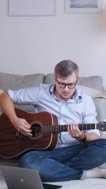 Dikey video. Gitar dersi. Mesafe dersi. Uzaktan eğitim. Akustik enstrüman çalan zeki bir adam ev içi fotokopi alanında dizüstü bilgisayarla yerde oturuyor..