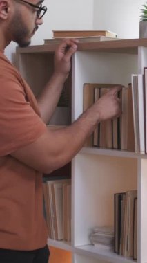 Dikey video. Kitap sever. Ev kütüphanesi. Hobi ediniyorum. Modern iç mekanlarda kitaplıktan okumak için edebiyat seçen gözlüklü zeki adam..