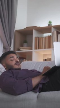 Dikey video. İnternet sohbeti. Sanal arama. Dijital bir eğlence. Rahat adam defter kullanarak sohbetten zevk alıyor. Modern ev oturma odasındaki rahat koltukta yatıyor. İçi fotokopi alanı dolu..