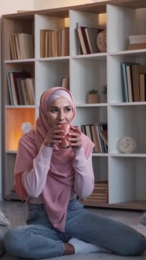 Dikey video. Ev kahvesi. Hafta sonu tatili. Kafiyeli ve kupalı mutlu kadın oturma odasında yerde sıcak içkinin keyfini çıkarıyor..