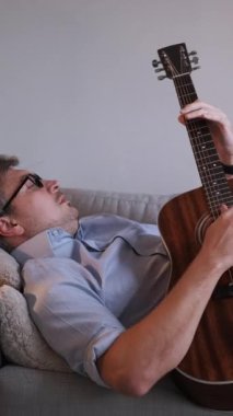 Dikey video. Müzisyen rahatla. Ev istirahatı. Huzurlu, sıradan, rüya gibi bir adam kanepeye uzanmış, akustik gitar çalıyor, iç mekânda melodi besteliyor..