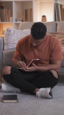 Dikey video. Ev ödevi. Hobi ediniyorum. Yaratıcı eğitim. Akıllı adam, elinde kitaplarla oturmuş modern oturma odasının iç kısımlarında notlar alıyor..