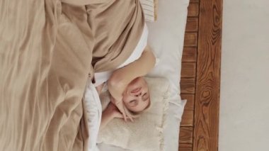 Dikey video. Sabah uyanması. Rahat bir uyku. Huzurlu, rahat bir kadın, yumuşak çarşaflı, rahat bir yatakta uzanıyor. Evinde yumuşak yastıklı bej bir battaniye var..