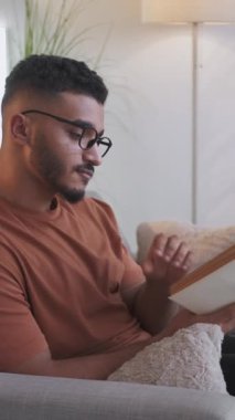 Dikey video. Boş vakitleri değerlendiriyorum. Ev istirahatı. Edebiyat hobisi. Gözlüklü, zeki, açık gözlü, modern oturma odasındaki boş odalı kanepenin sayfalarını çeviren bir adam..