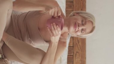 Dikey video. Kahve gevşedi. Hafta sonu istirahati. Ev atmosferi. Mutlu rüya gibi kadın bir fincan çay içiyor. Pazar keyfine bakıyor. Işıl ışıl iç mekanda oturuyor..