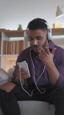 Dikey video. Sanal sohbet. Çevrimiçi çağrı. Dijital iletişim. Kulaklık takan kendine güvenen bir adam modern iç mekandaki rahat koltukta telefonla konuşuyor..