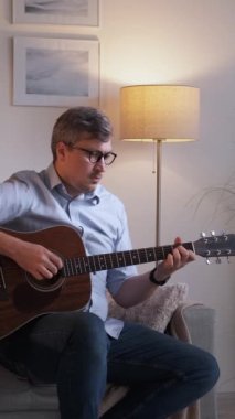Dikey video. Müzik bestecisi. İlham verici gitarist. Kanepede oturup akustik enstrüman çalan duygusal bir adam evde oturma odasında yaratıcı bir hobiyle eğleniyor..