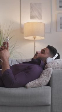 Dikey video. Sanal ev araması. Çevrimiçi sohbet. Mesafe iletişimi. Kulaklıklı rahat adam telefonda konuşuyor. Modern oturma odasının iç tarafındaki rahat koltukta uzanıyor..