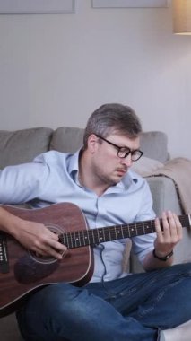 Dikey video. Müzik öğrenme. Akustik ses. Zeki, ilham verici bir adam klasik gitar çalarak ev fotokopi odasının içindeki oturma odasında melodinin tadını çıkarıyor..