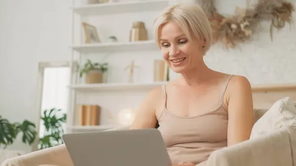 stock image Virtual connection. Online communication. Excited smiling woman talking at video conference on laptop sitting in armchair light living room home interior.