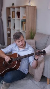 Dikey video. Müzisyen hobisi. İlham verici oyuncu. Rahatlamış yetenekli bir adam yerde oturmuş akustik gitar akorlarının keyfini çıkarıyor hafta sonları evde, oturma odasında keyif çatıyor..
