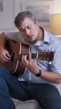 Dikey video. Müzikal hobi. Gitar alıştırması. Mutlu, gülümseyen adam evde oturup telli çalgılar çalıyor. Oturma odasının iç kısımlarında akorları öğreniyor..