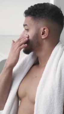 Dikey video. Cilt nemlendirici. Yüz bakımı. Sabah rutini. Havluyla banyodan sonra yüz kremi süren kendinden emin bir adam..