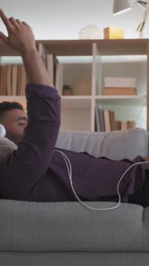 Dikey video. Müzik enerjisi. Ev tatili. Pozitif enerji. Oturma odasındaki kanepede oturup kulaklıkla müzik dinlemekten keyif alan rahat adam..
